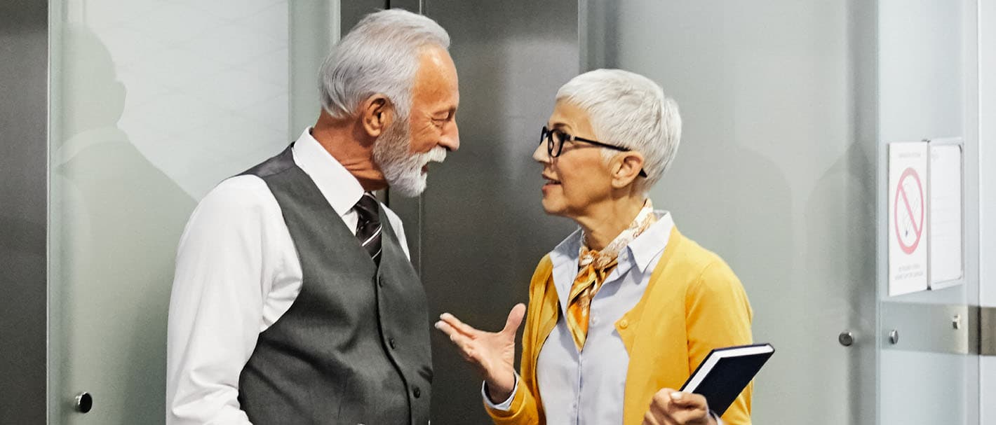 Two older business people, a man and a woman, are standing in front of an elevator in an office building and talking.